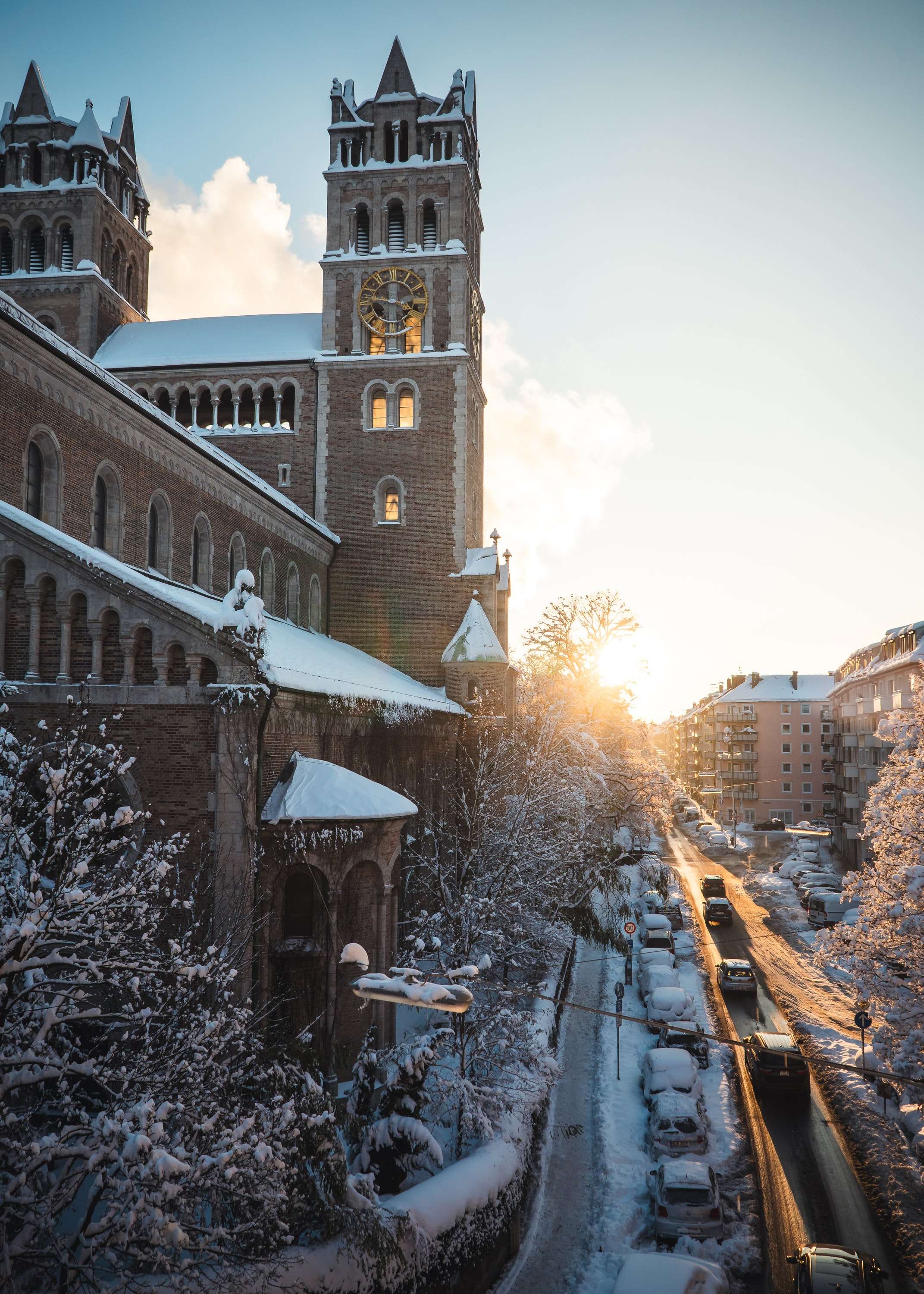 The brick towers of St. Benno's church in Munich above a snow-covered street lined with parked cars, the low sun shining down the road.