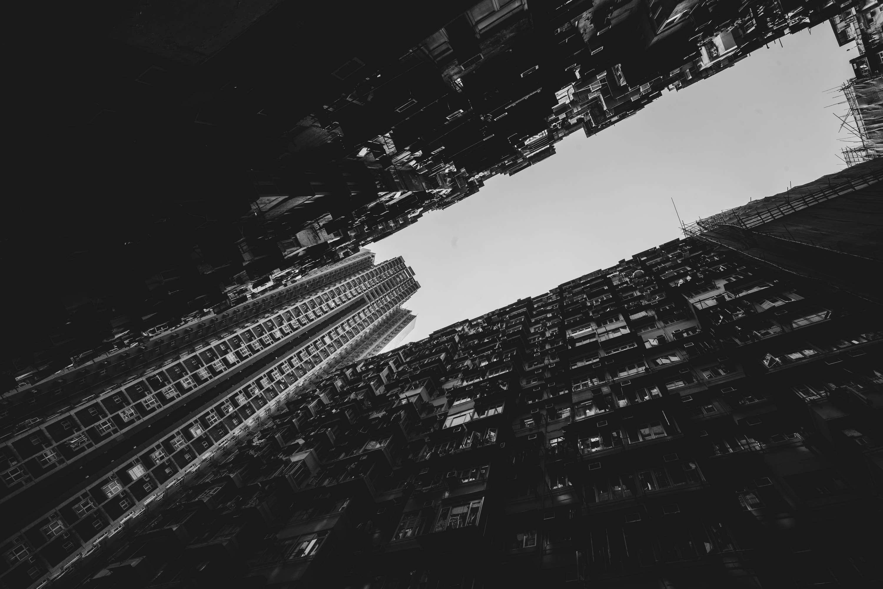 Black and white: looking straight up between towering residential blocks, their windows and balconies converging on a thin strip of grey sky.
