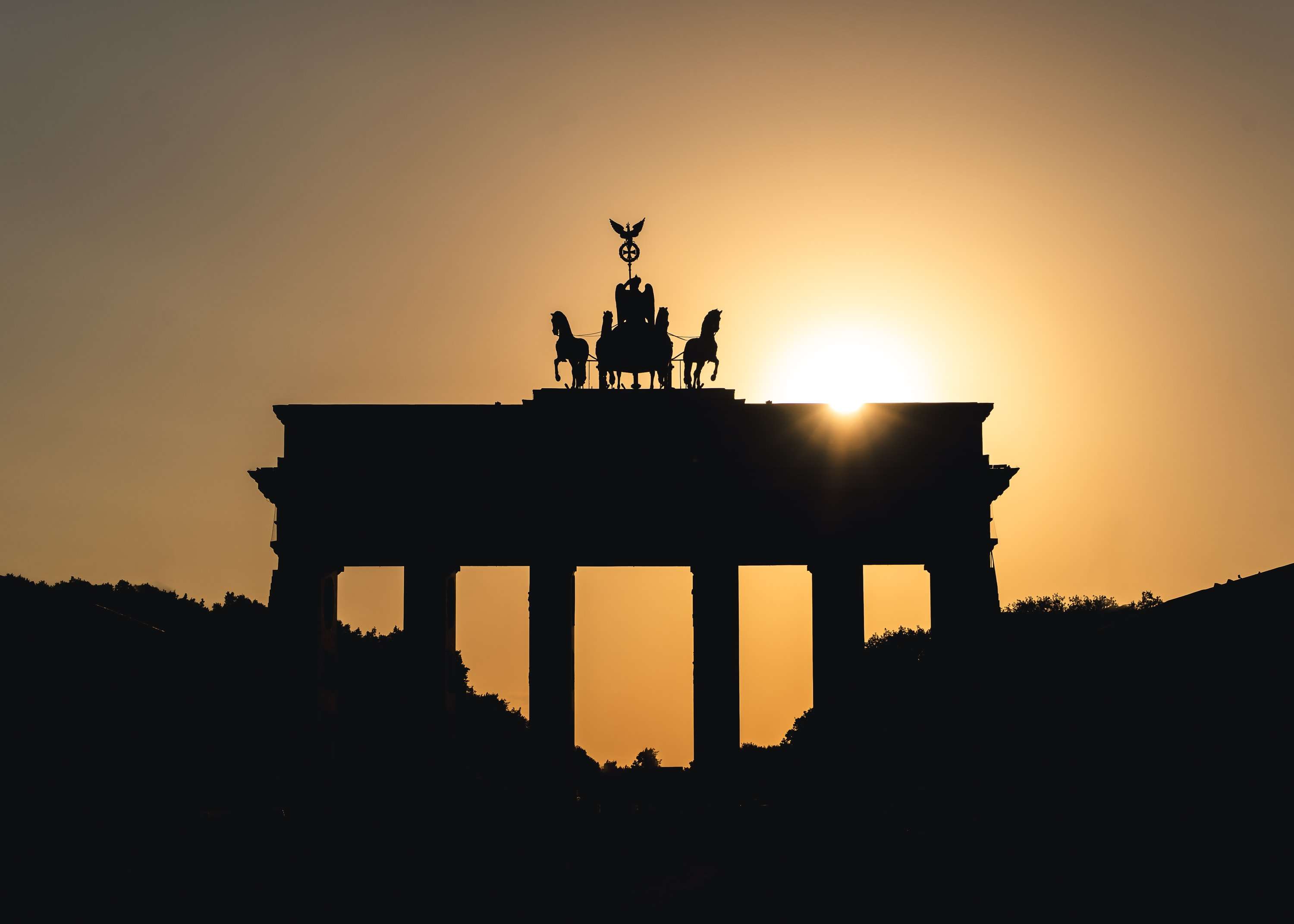The Brandenburg Gate in black silhouette against an orange sky, the sun setting just behind the quadriga on top.