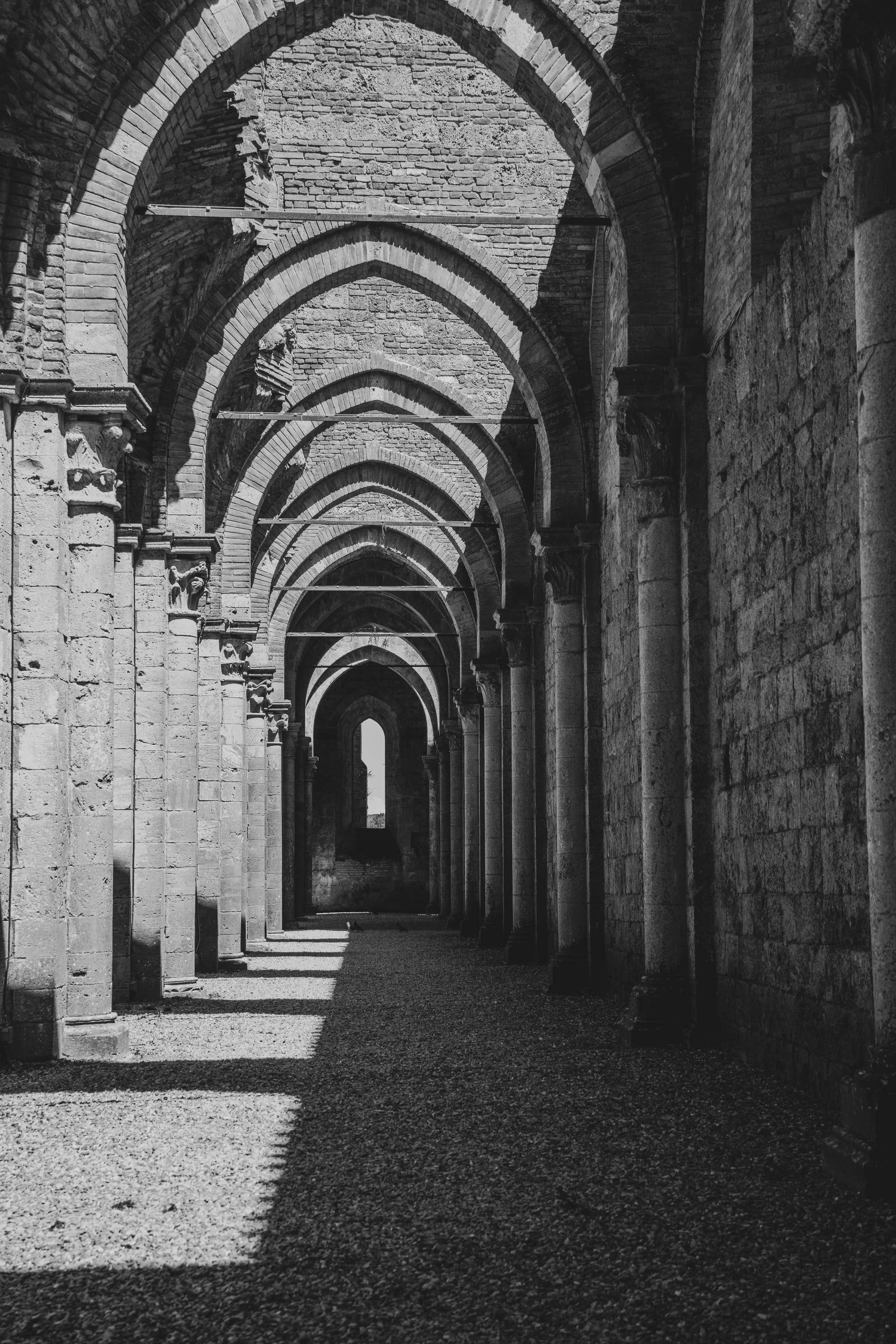 Black and white: the roofless nave of San Galgano Abbey, a long row of pointed stone arches with sunlight falling in patches on the gravel floor.
