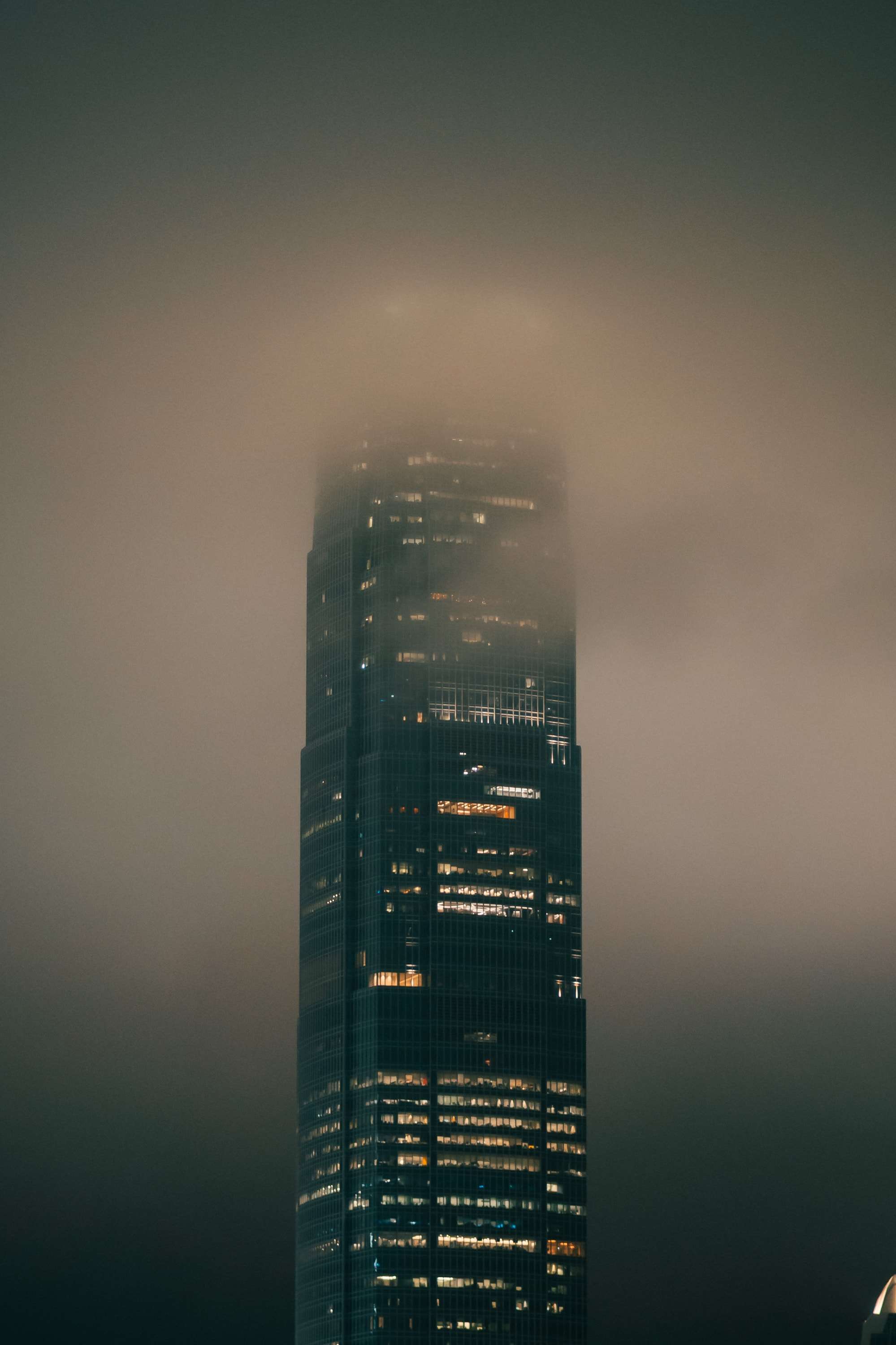 The International Finance Centre tower, windows lit, rises into thick fog that hides its top, against a dark green sky.
