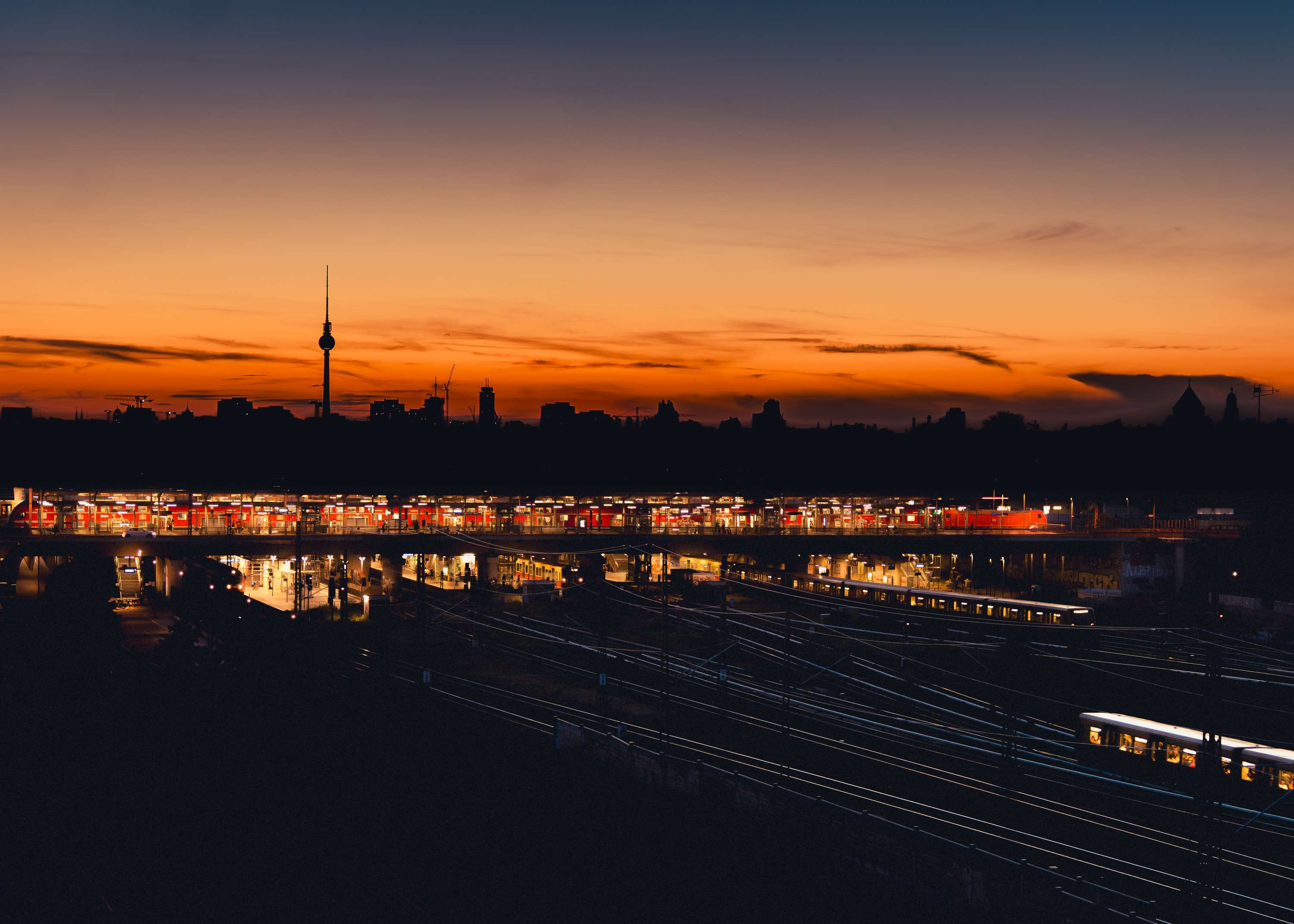The lit platforms of Warschauer Straße station after sunset, trains on the tracks below, with the Fernsehturm small on a dark skyline under an orange band of sky.