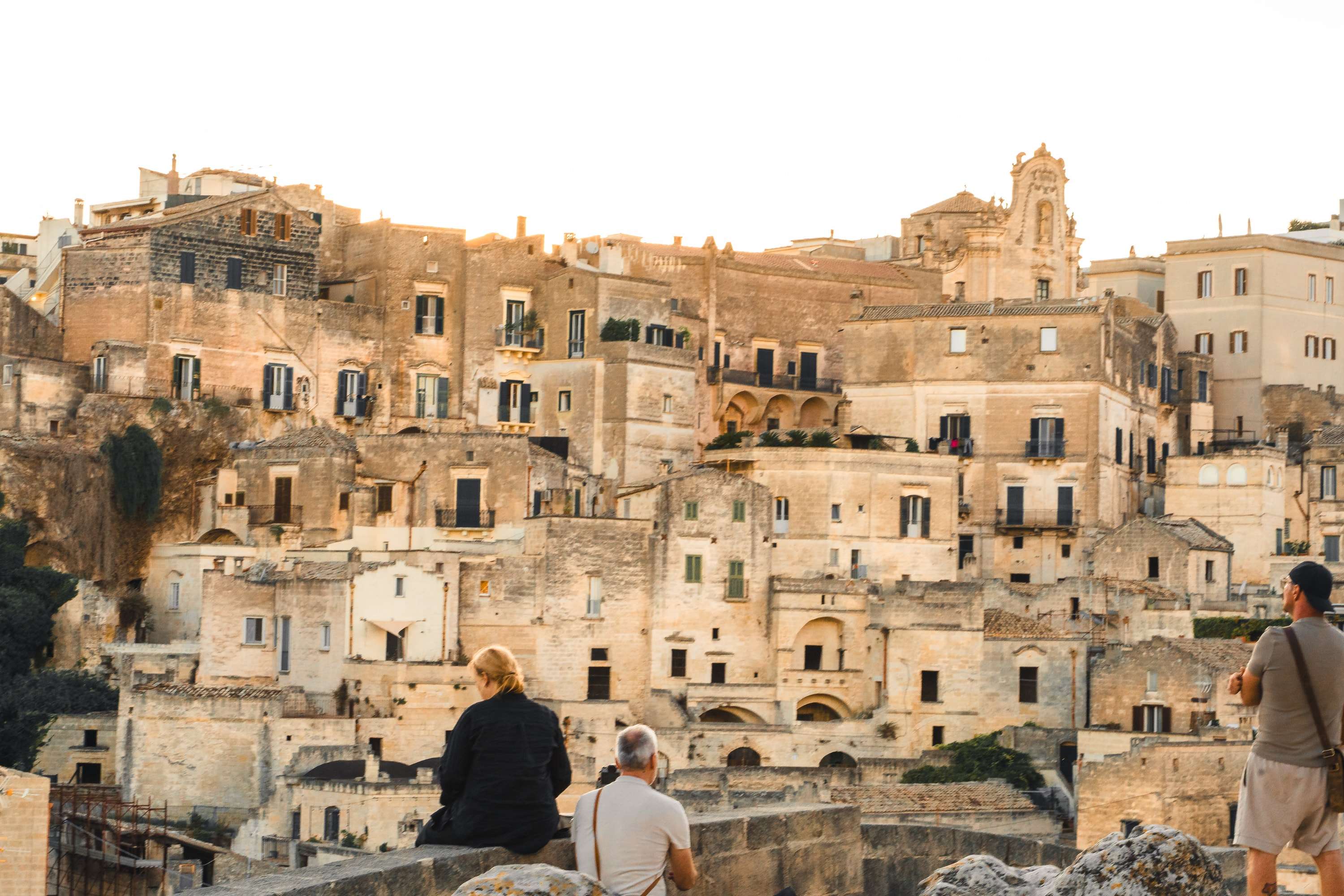 Three people sit and stand on a stone wall looking across the pale stone houses of the Sassi di Matera, stacked up the hillside in evening light.