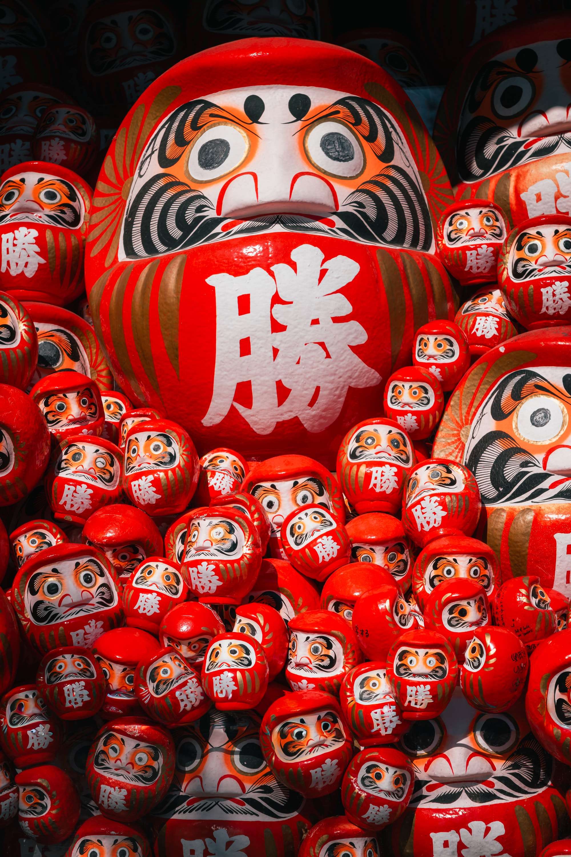 Hundreds of red daruma dolls marked with the character for victory are piled together, one large daruma in the centre looking straight at the camera.