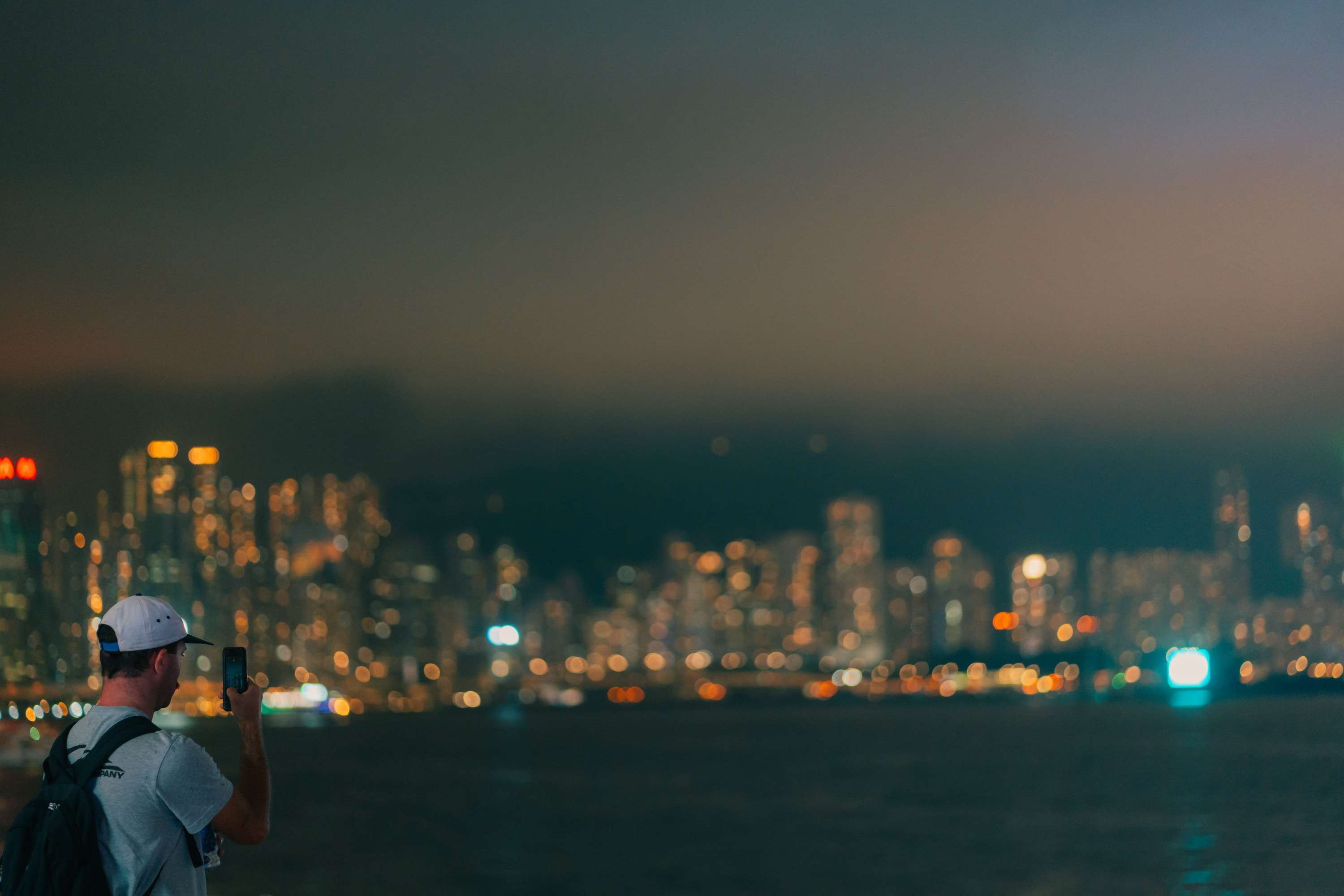 A man in a white cap and backpack holds up his phone to photograph the Hong Kong skyline, which is blurred into points of light across the harbour.