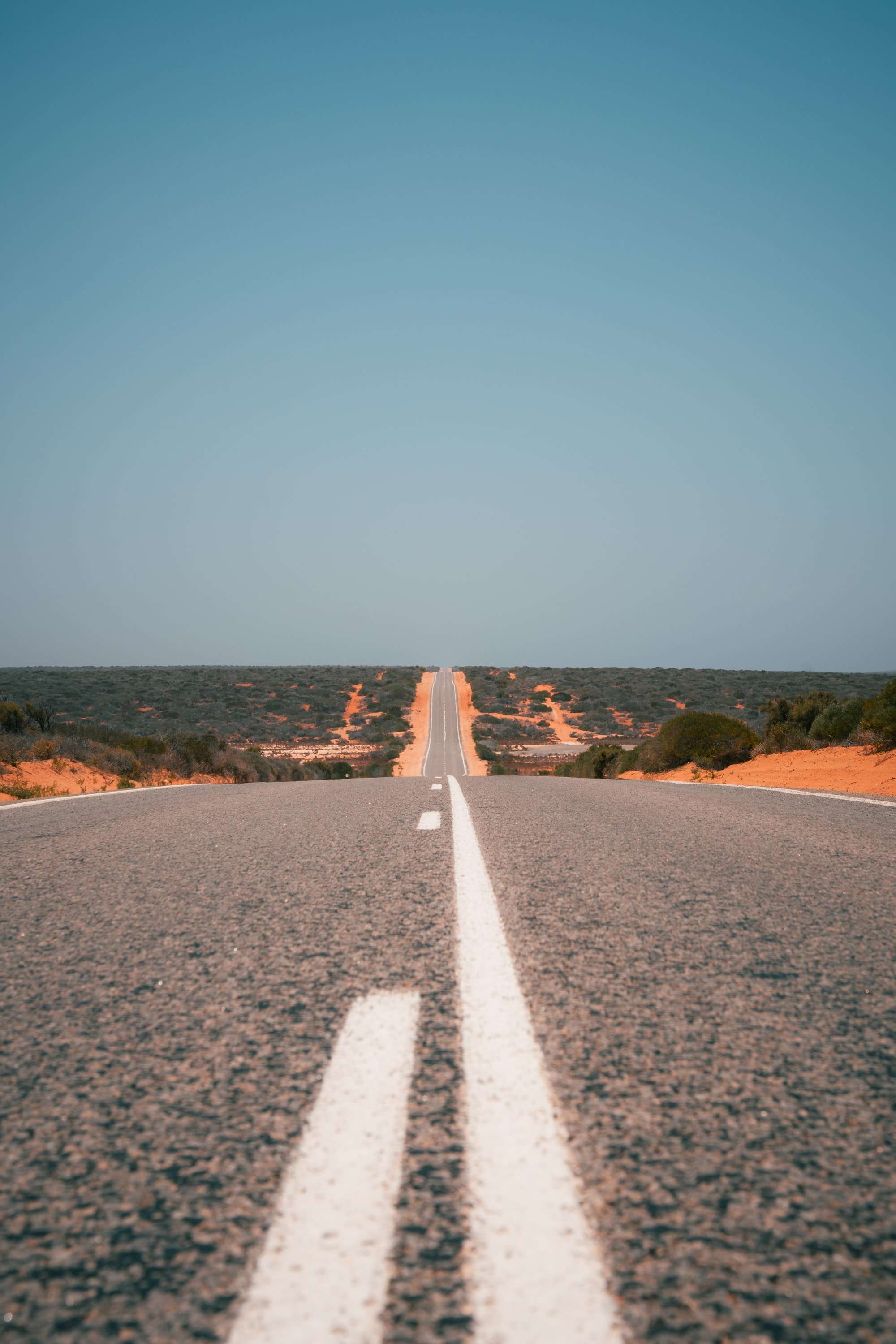 A straight two-lane road runs to the horizon between low scrub and red sand dunes, the white centre line leading up from the foreground.