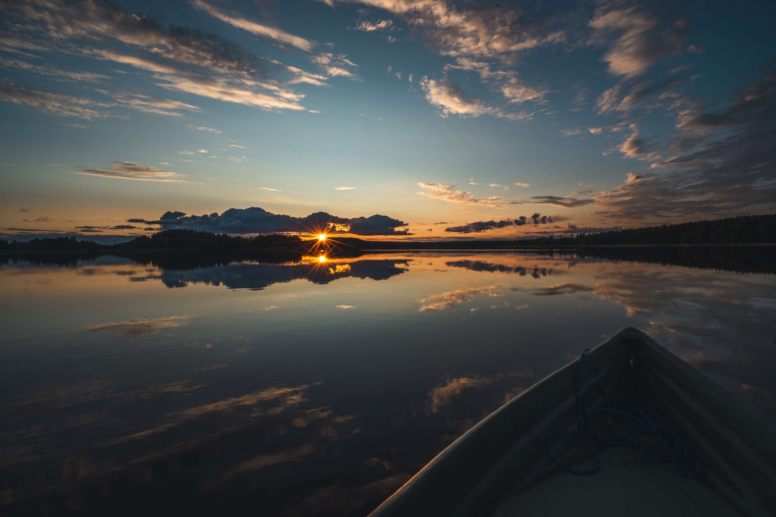 The bow of a small boat points across a still lake at sunset, the sun setting behind a wooded island and the clouds reflected in the water.