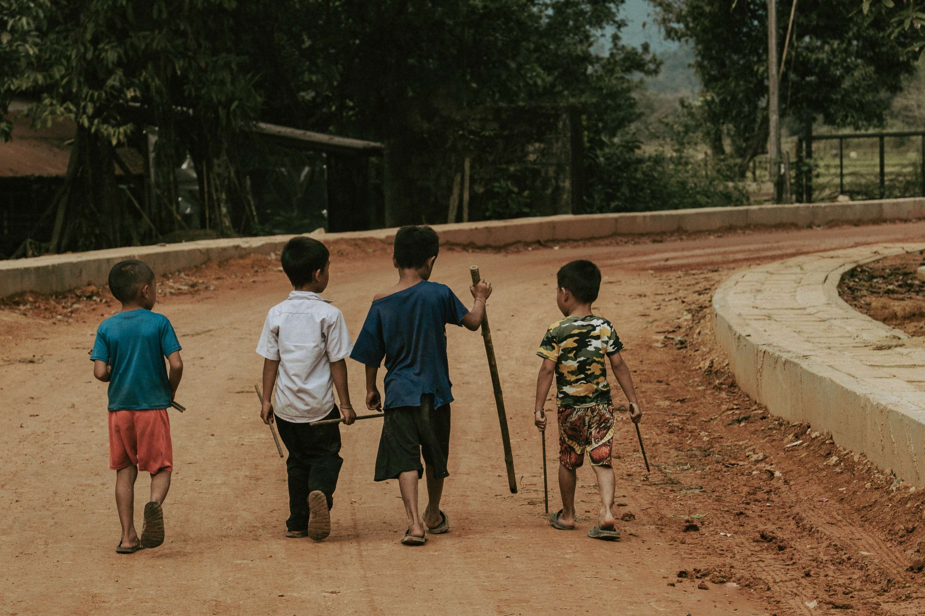 Four young boys walk away from the camera down a red dirt road, each carrying a long stick, one in a white school shirt.