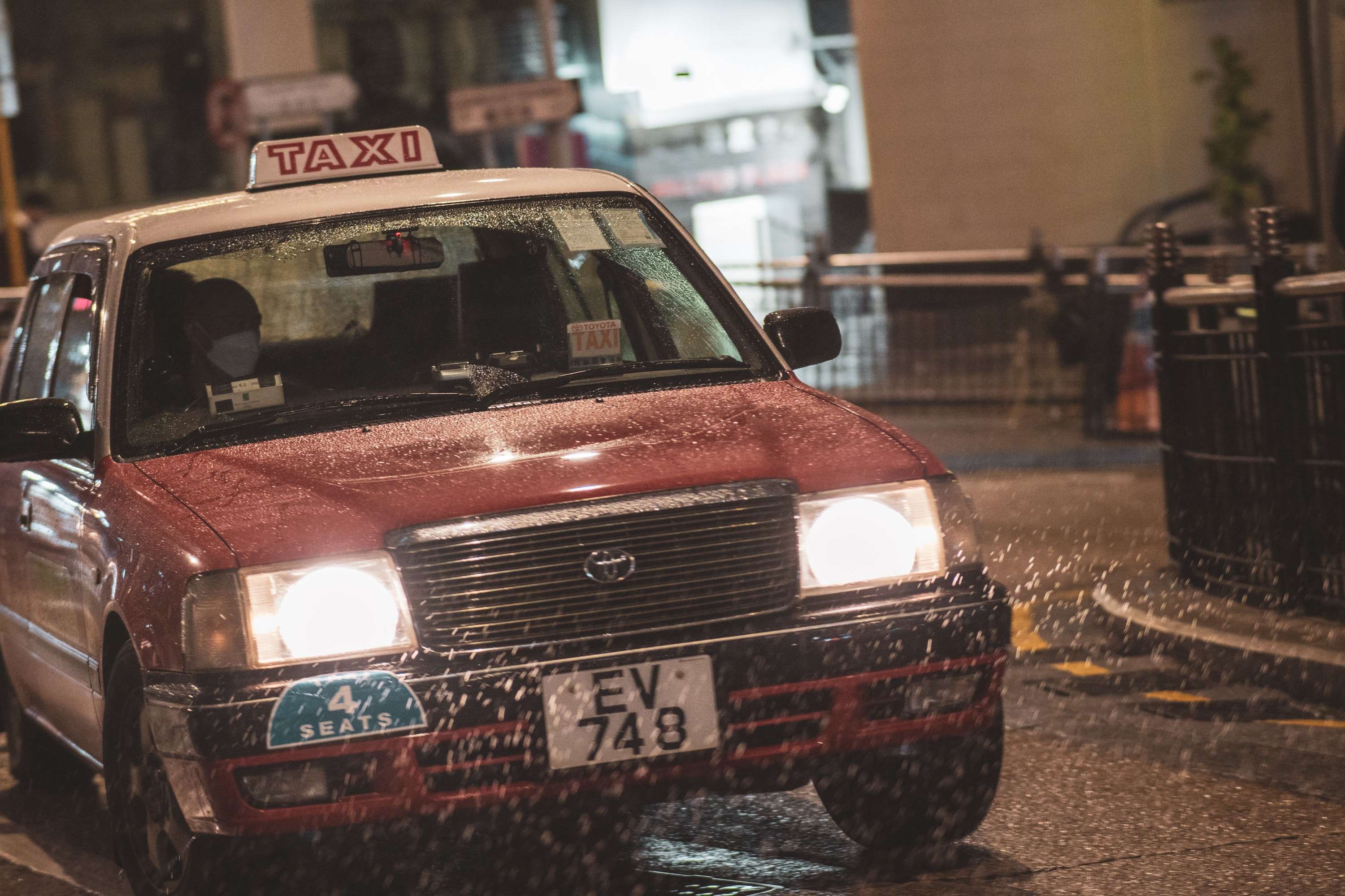 A red Hong Kong taxi with its headlights on turns through a wet street at night, rain bouncing off its bonnet.