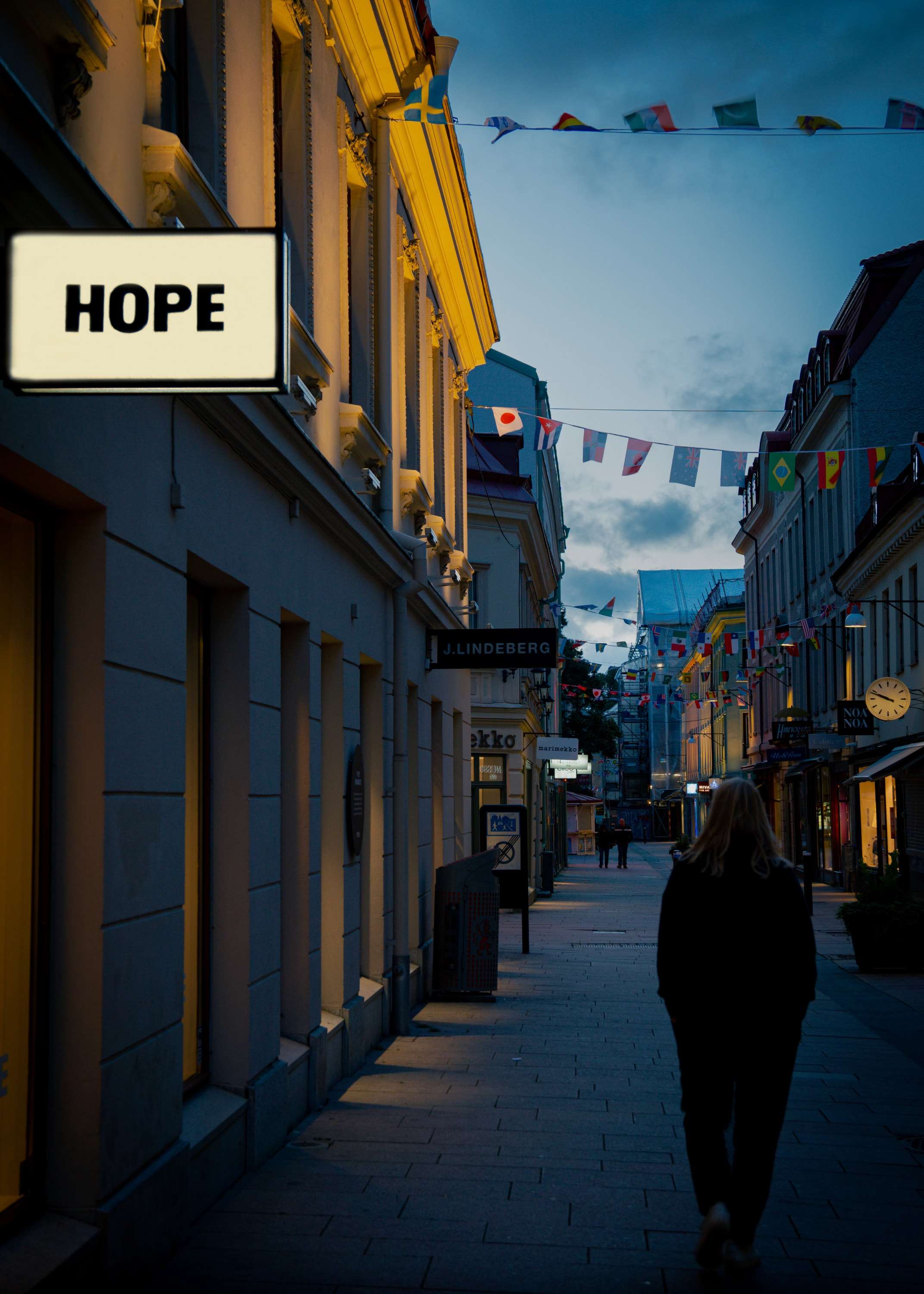 A woman walks away down an evening street past a lit sign reading HOPE, with strings of flags hung between the buildings.
