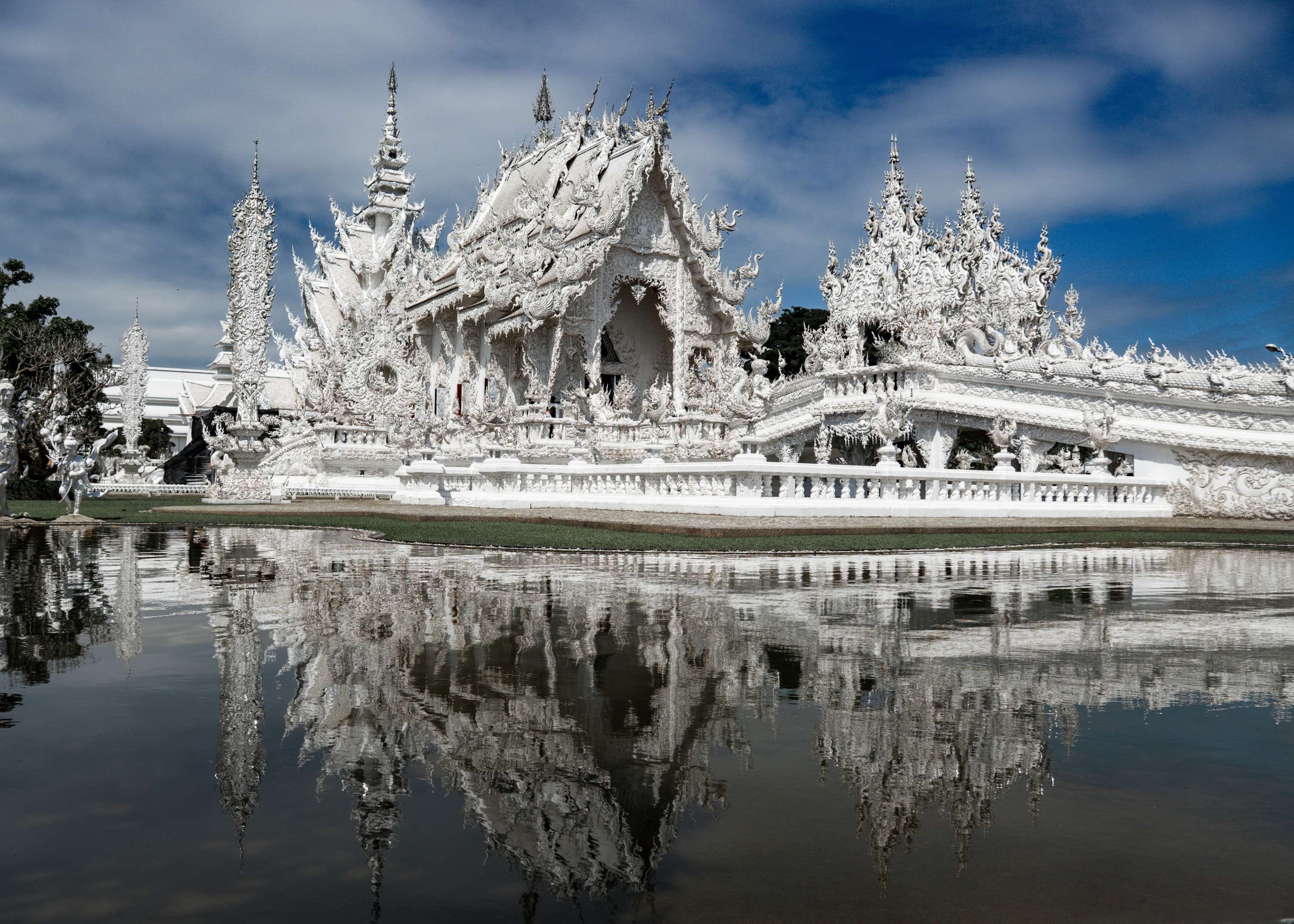 The all-white Wat Rong Khun temple with its spiked roofs and carved balustrades, reflected almost perfectly in the pond in front of it under a blue sky.