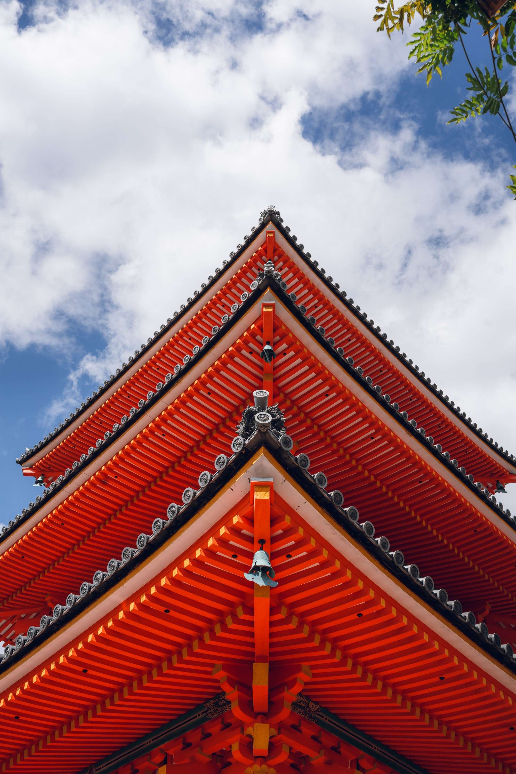 The pointed vermilion roofs of a three-storey pagoda rise into a blue sky with white clouds, a small bell hanging below the lowest eave.