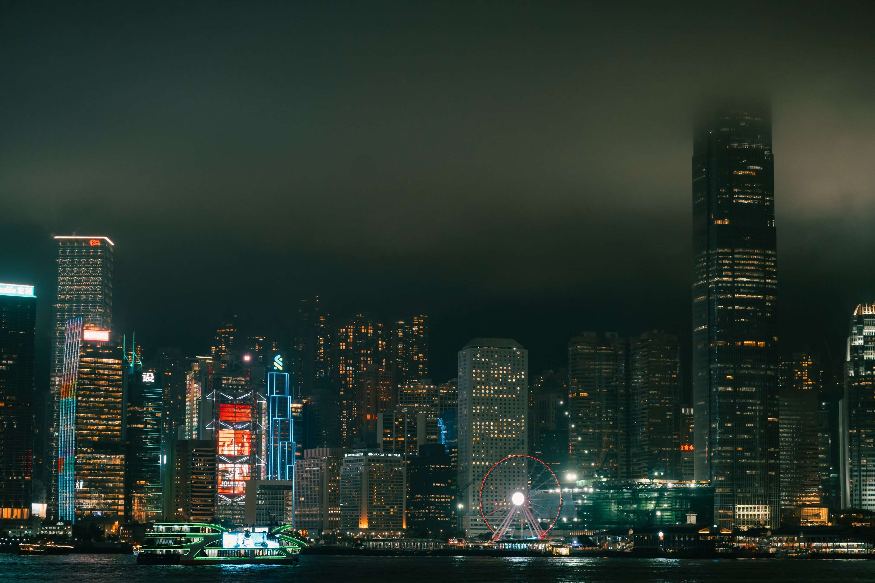 The Hong Kong Island skyline at night across Victoria Harbour, low cloud swallowing the top of the IFC tower, a Ferris wheel lit red at the waterfront.
