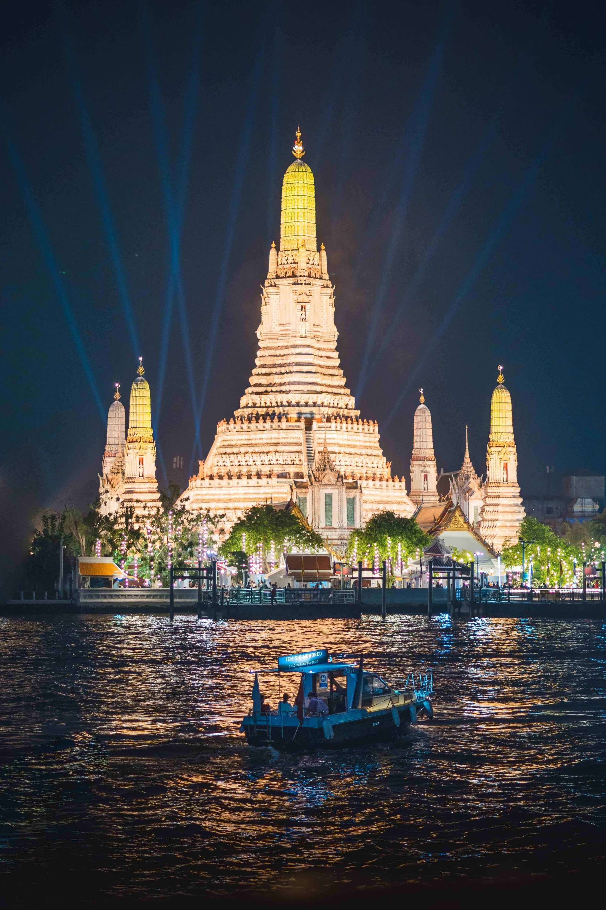 The lit central prang of Wat Arun at night, reflected in the Chao Phraya river with a boat passing in front.