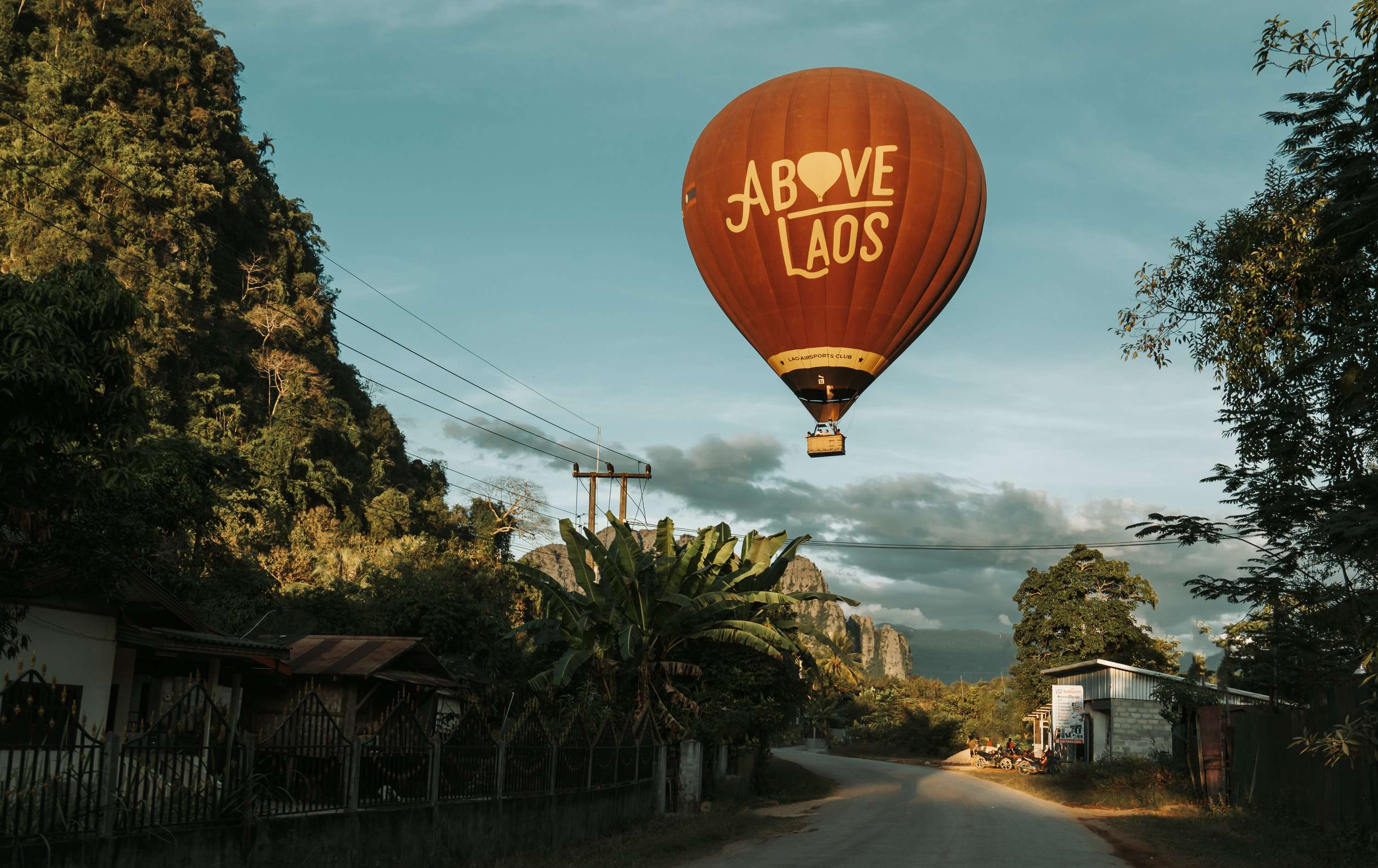 An orange hot-air balloon printed with the words Above Laos floats over a village road lined with trees, houses and power lines, limestone hills behind.
