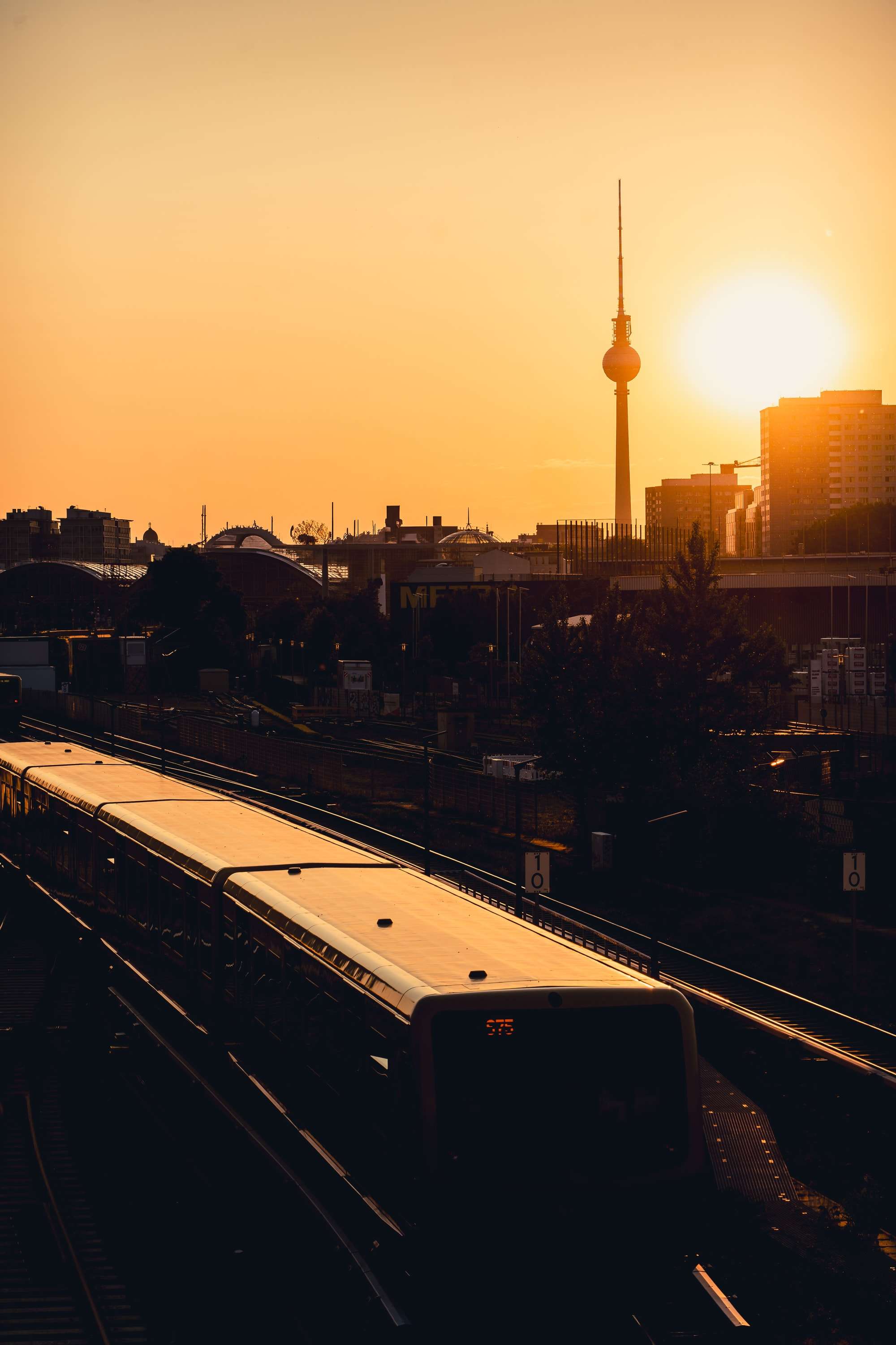 A Berlin S-Bahn train runs along the tracks in the evening, its roof lit orange by the setting sun, with the Fernsehturm on the skyline beside the sun.